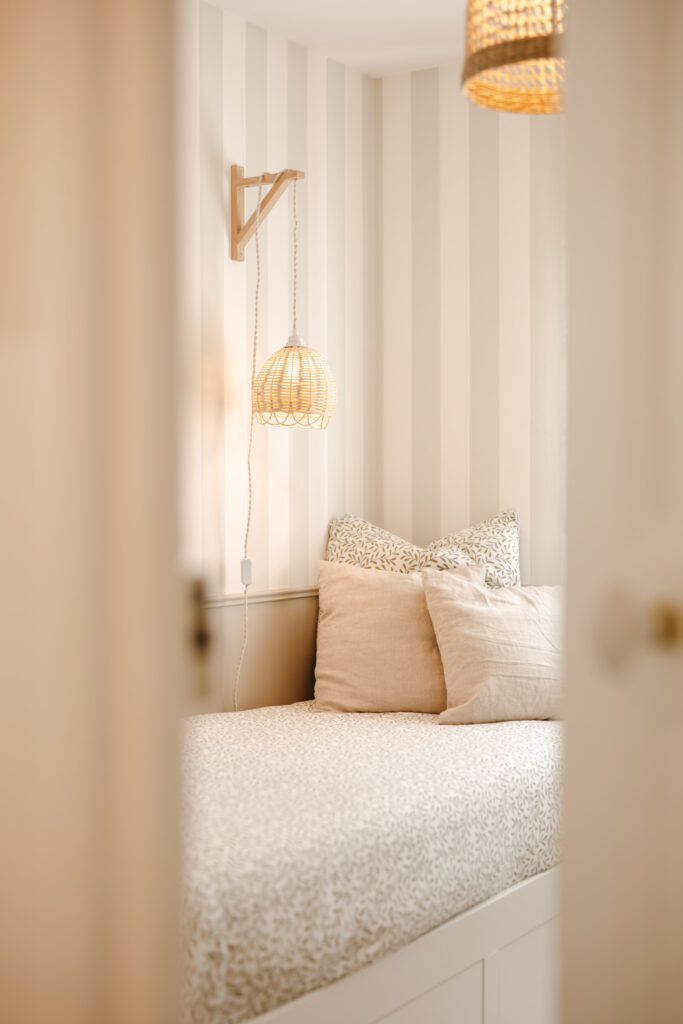 Lit simple avec coussins beiges et imprimés végétaux, contre un mur à rayures verticales vert et blanc, suspension en rotin, décoré par Maison Rivage à Porspoder.